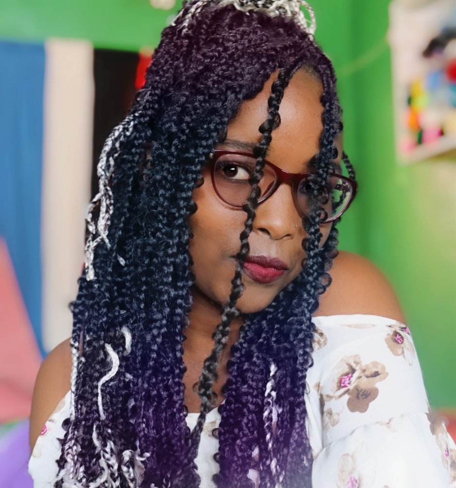 Mary Wanjiru, wearing maroon glasses and an off-shoulder white floral top, with long curly braids styled with white highlights, posing confidently indoors against a colorful background